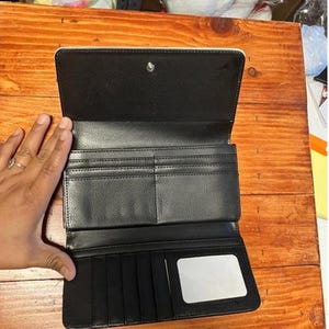 May include: Black bi-fold wallet with multiple card slots, bill compartments, and a clear ID window. The wallet is open, revealing its interior. The exterior is black with a silver snap closure.