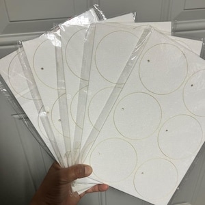 May include: A hand holding several packages of white paper sheets. Each sheet has a series of gold-colored circular outlines and small holes. The sheets are packaged in clear plastic.