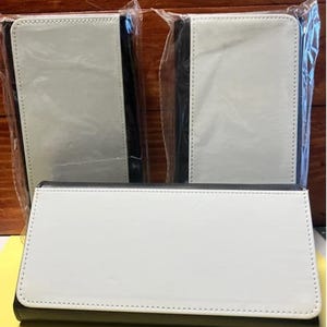 May include: Three white and black wallets, each in a protective plastic sleeve. The wallets have a white front with a black border and a stitched edge. The wallets are rectangular and appear to be made of a smooth material.