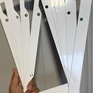 May include: A stack of white license plate frames. Each frame is rectangular with rounded corners and pre-drilled holes for mounting. The frames are held in a hand against a neutral background.