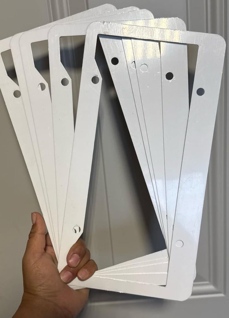 May include: A stack of white license plate frames. Each frame is rectangular with rounded corners and pre-drilled holes for mounting. The frames are held in a hand against a neutral background.