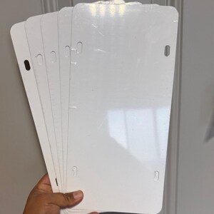 May include: A stack of blank, white license plates. Each plate has rounded corners and pre-drilled holes for mounting. The plates are held in a hand, with the white surface reflecting light. The background is a light gray door.