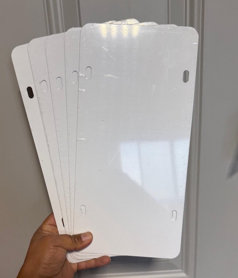 May include: A stack of blank, white license plates. Each plate has rounded corners and pre-drilled holes for mounting. The plates are held in a hand, with the white surface reflecting light. The background is a light gray door.