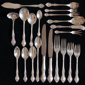 May include: An assortment of antique silverware, including spoons, forks, knives, and a ladle, arranged on a black surface. The silverware has ornate, detailed handles and a shiny, reflective surface. The collection includes various sizes and styles.