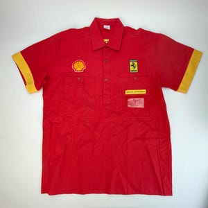 Puede incluir: Camisa roja de manga corta con detalles amarillos en las mangas. La camisa presenta el logotipo de Shell, un emblema de Ferrari y un parche que dice "SERVICO ANTICENDIOS". Tiene dos bolsillos en el pecho y cuello abotonado.