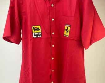 Può includere: Camicia rossa brillante a maniche corte con colletto button-down. Presenta due toppe: una con il logo Agip e l'altra con il logo Ferrari. La camicia è appesa a una gruccia di legno.
