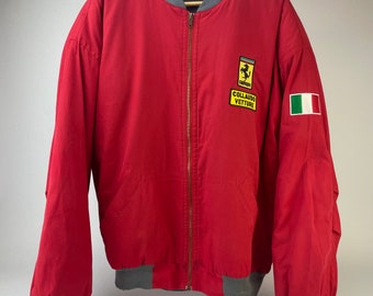 May include: A red bomber jacket with a gray zipper and ribbed cuffs. The jacket has a yellow patch with the Ferrari logo and the text "COLLAUDO VETTURE". The jacket also has a small Italian flag patch on the left sleeve.