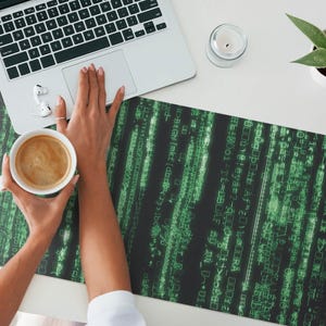 Può includere: Un tappetino per mouse nero e verde con un motivo di codice verde su sfondo nero. Una persona tiene una tazza di caffè bianca con un liquido marrone all'interno.