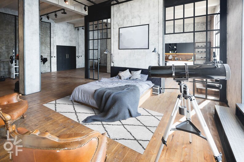 May include: A modern loft bedroom with a gray bed, a white and black patterned rug, two brown leather chairs, and a telescope on a tripod.