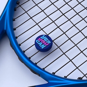 May include: A blue tennis racket with black strings and a small, round, blue button. The button has the words "YES YOU CAN" in pink and blue lettering with a smiling face graphic. The racket is angled diagonally.