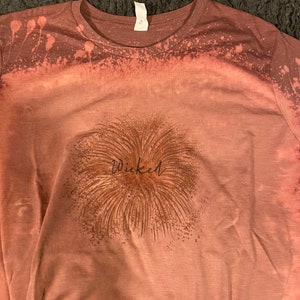 Peut inclure: Un t-shirt marron avec un motif éclaboussé de javel. Le t-shirt a un graphique marron d'un feu d'artifice avec le mot "Wicked" écrit en cursive noire.