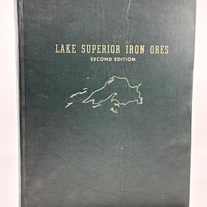 May include: A dark green hardcover book titled Lake Superior Iron Ores Second Edition with a gold outline map of the lake on the front cover. A wooden ruler is positioned at the bottom, showing the book is approximately 11 inches in height.