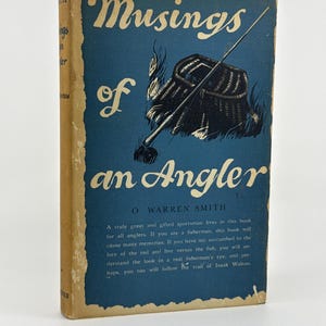 May include: A hardcover book titled Musings of an Angler by O. Warren Smith. The blue cover features a cream-colored illustration of a fishing creel and rod. The text includes the title, author name, and a descriptive paragraph on the front.