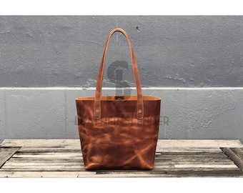 Peut inclure: Un sac cabas en cuir brun avec deux anses et une fermeture à bouton pression. Le sac est en cuir vieilli et a un aspect rustique.