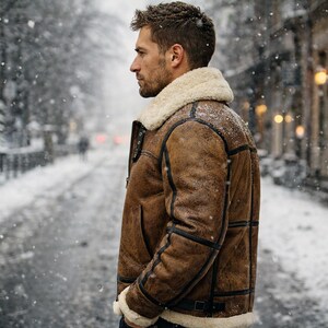 May include: A brown shearling jacket with a cream-colored shearling collar and cuffs. The jacket features black accents and a buckled belt. The person is standing on a snow-covered street, with falling snow and a blurred background.