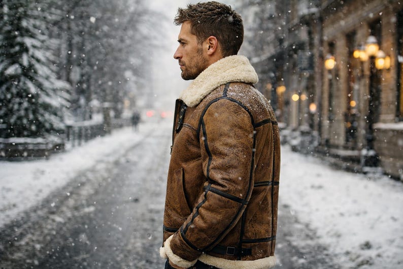 May include: A brown shearling jacket with a cream-colored shearling collar and cuffs. The jacket features black accents and a buckled belt. The person is standing on a snow-covered street, with falling snow and a blurred background.