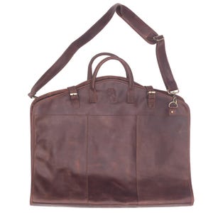 May include: A brown leather garment bag with a shoulder strap and a top handle. The bag has a flap closure with a buckle and a zippered pocket on the front.