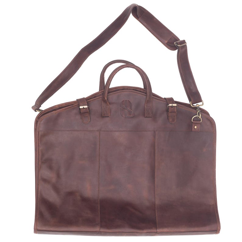 May include: A brown leather garment bag with a shoulder strap and a top handle. The bag has a flap closure with a buckle and a zippered pocket on the front.