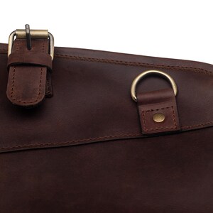 May include: Close-up of a brown leather bag with a brass buckle and a metal ring. The bag has a stitched seam and a smooth, textured surface.
