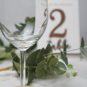 Puede incluir: Una copa de vino de vidrio transparente se encuentra sobre un posavasos de madera con la palabra "Michelle" grabada. La copa est&aacute; delante de una tarjeta blanca con el n&uacute;mero "2" y la palabra "mesa" escrita en marr&oacute;n.