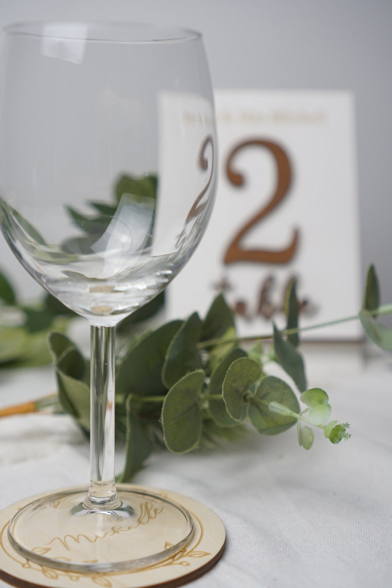 Puede incluir: Una copa de vino de vidrio transparente se encuentra sobre un posavasos de madera con la palabra "Michelle" grabada. La copa est&aacute; delante de una tarjeta blanca con el n&uacute;mero "2" y la palabra "mesa" escrita en marr&oacute;n.