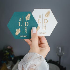 Può includere: Due sottobicchieri esagonali, uno verde acqua e uno bianco, sono mostrati. Ogni sottobicchiere presenta scritte dorate con i nomi "Liz" e "Daniel", la data "10.7.2025" e un motivo a felce. I sottobicchieri sono probabilmente per un matrimonio.