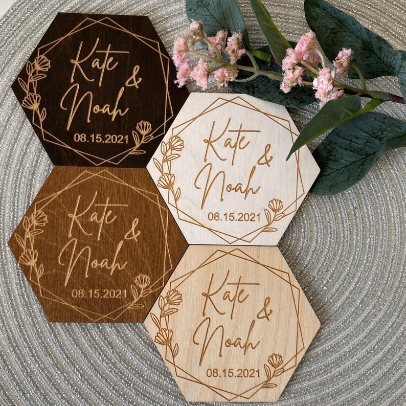Puede incluir: Cuatro posavasos hexagonales de madera con un dise&ntilde;o floral y el texto "Kate & Noah 08.15.2021" grabado en cada uno. Dos posavasos son de color marr&oacute;n claro y dos son de color marr&oacute;n oscuro.