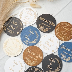 Puede incluir: Un conjunto de diez posavasos redondos de madera con dise&ntilde;os florales grabados y nombres. Los posavasos son de varios colores, incluyendo blanco, marr&oacute;n, negro y azul. El texto en los posavasos incluye nombres como "Julie y Chris", "Ashley y Josh" y la fecha del evento.