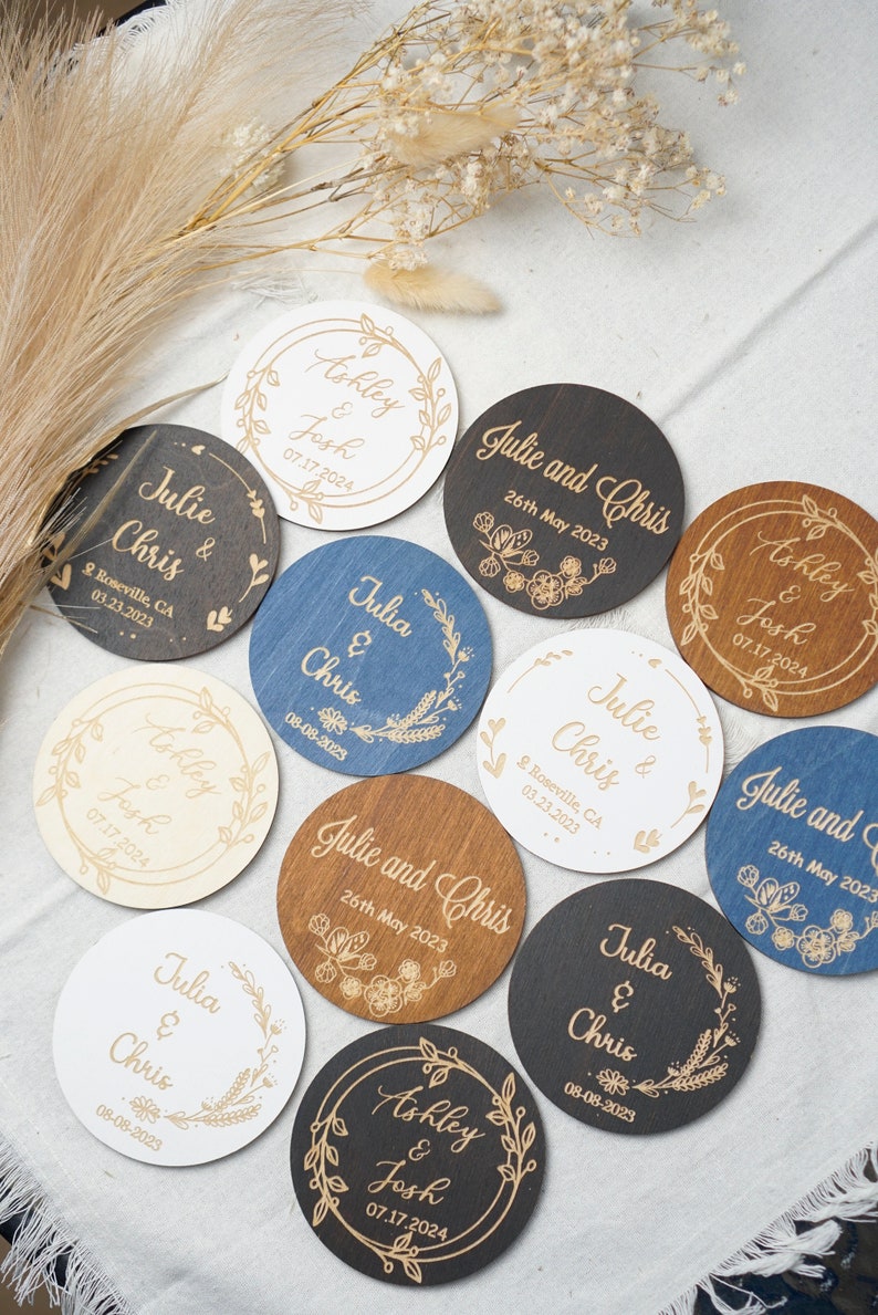 Puede incluir: Un conjunto de diez posavasos redondos de madera con dise&ntilde;os florales grabados y nombres. Los posavasos son de varios colores, incluyendo blanco, marr&oacute;n, negro y azul. El texto en los posavasos incluye nombres como "Julie y Chris", "Ashley y Josh" y la fecha del evento.