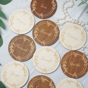 Puede incluir: Diez tarjetas de lugar redondas de madera con un dise&ntilde;o floral y nombres escritos en cursiva. Los nombres son: Tori, Maria, Dawide, Natalie, Hannah, Sarah, Yvette, Diego, Sandra y Michelle.