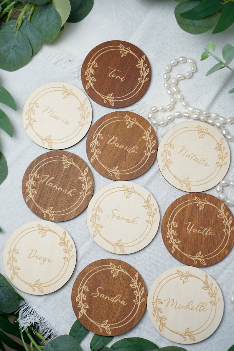 Puede incluir: Diez tarjetas de lugar redondas de madera con un dise&ntilde;o floral y nombres escritos en cursiva. Los nombres son: Tori, Maria, Dawide, Natalie, Hannah, Sarah, Yvette, Diego, Sandra y Michelle.
