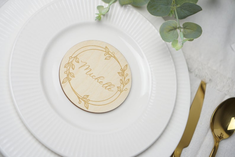 Puede incluir: Una tarjeta de lugar redonda de madera con un dise&ntilde;o floral y el nombre "Mickelle" grabado en el centro.