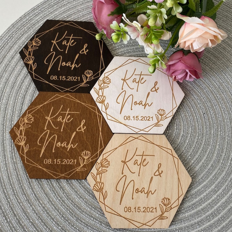 Puede incluir: Cuatro posavasos hexagonales de madera con un dise&ntilde;o geom&eacute;trico dorado y detalles florales. Cada posavasos tiene el texto "Kate & Noah" y la fecha "08.15.2021".
