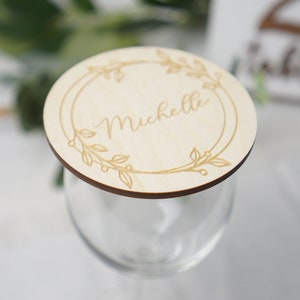Puede incluir: Una tarjeta de lugar redonda de madera con un dise&ntilde;o floral y el nombre "Michelle" grabado en el centro. La tarjeta de lugar est&aacute; colocada encima de una copa de vino transparente.