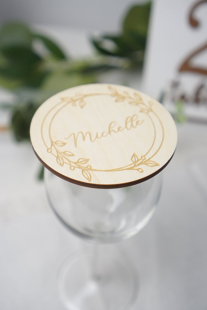 Puede incluir: Una tarjeta de lugar redonda de madera con un dise&ntilde;o floral y el nombre "Michelle" grabado en el centro. La tarjeta de lugar est&aacute; colocada encima de una copa de vino transparente.