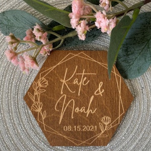 Puede incluir: Un letrero hexagonal de madera con un marco geom&eacute;trico y detalles florales. El letrero est&aacute; grabado con los nombres "Kate & Noah" y la fecha "08.15.2021".