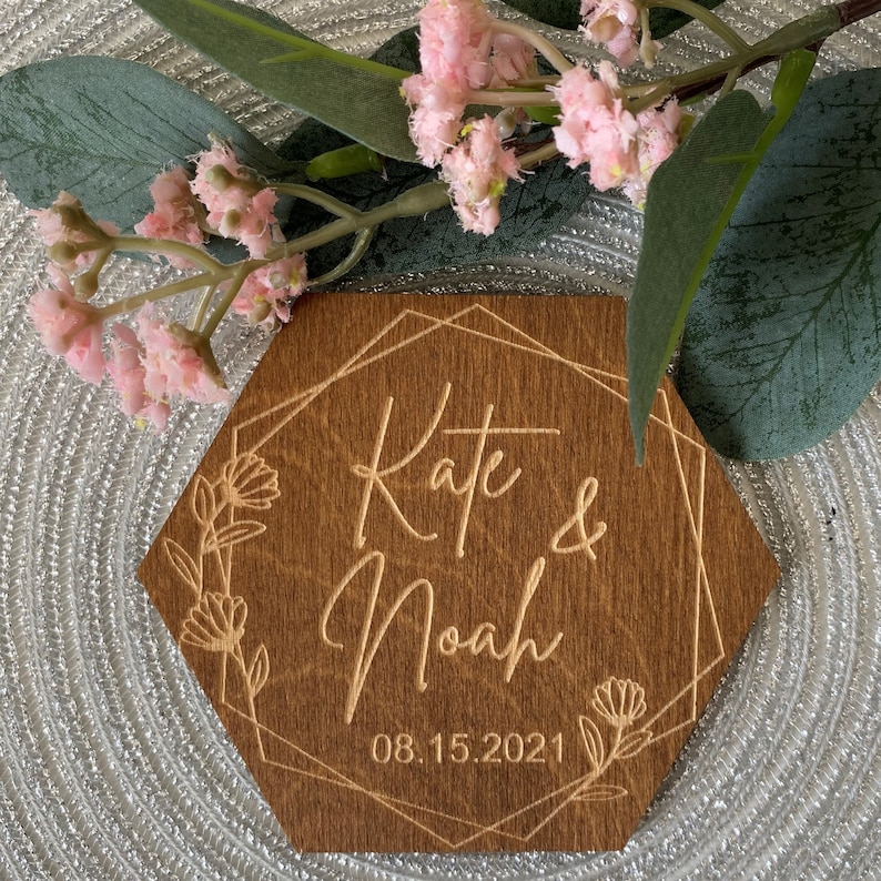 Puede incluir: Un letrero hexagonal de madera con un marco geom&eacute;trico y detalles florales. El letrero est&aacute; grabado con los nombres "Kate & Noah" y la fecha "08.15.2021".