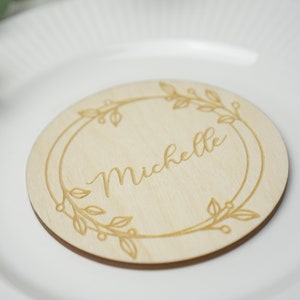 Puede incluir: Una tarjeta de lugar redonda de madera con un dise&ntilde;o floral grabado alrededor del borde. El nombre "Michelle" est&aacute; grabado en el centro.