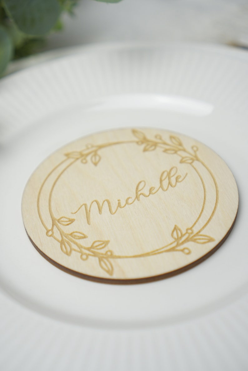 Puede incluir: Una tarjeta de lugar redonda de madera con un dise&ntilde;o floral grabado alrededor del borde. El nombre "Michelle" est&aacute; grabado en el centro.