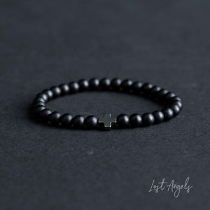 May include: A black beaded bracelet with a small silver cross charm. The bracelet is made of matte black beads and has a simple, minimalist design.