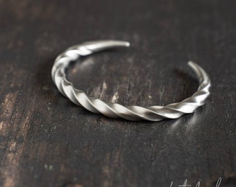 May include: A silver cuff bracelet with a twisted rope design. The open-ended bracelet is displayed on a dark wooden surface. The bracelet has a matte finish and is slightly curved.