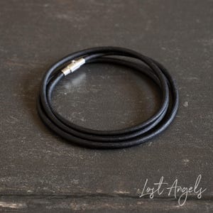 May include: A black leather cord necklace featuring a smooth, rounded texture and a silver-toned cylindrical magnetic clasp. The cord is coiled into a circular shape, showcasing its flexible design and the metallic finish of the fastener.
