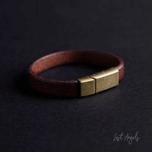 Puede incluir: Una pulsera de cuero marrón con un cierre rectangular de color dorado. La pulsera tiene un diseño de banda simple, con un color marrón oscuro. El cierre tiene forma rectangular. La pulsera está sobre una superficie negra.