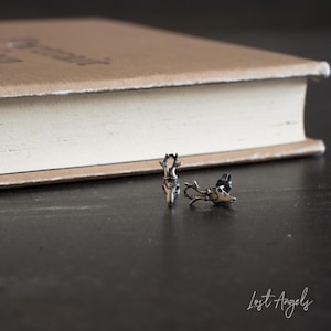 Peut inclure: Une paire de boucles d'oreilles en argent en forme de crâne de cerf avec des bois. Les boucles d'oreilles sont sur une surface sombre avec un livre en arrière-plan.