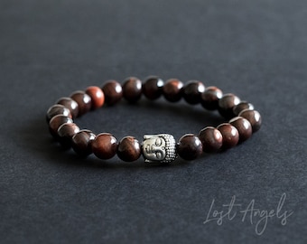 Puede incluir: Un brazalete de cuentas marrones con un dije de cabeza de Buda plateado. Las cuentas son una mezcla de marrón oscuro y rojo.