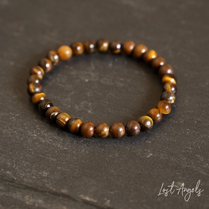 May include: A circular bracelet composed of polished tiger's eye beads in shades of golden brown and dark brown. The beads are strung together in a continuous loop. The text "Lost Angels" is visible in white script in the bottom right corner.