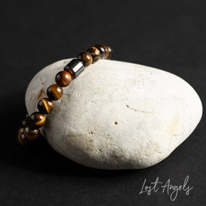 Puede incluir: Un brazalete de cuentas de ojo de tigre marrón y dorado con un acento de metal plateado. El brazalete está apoyado en una roca blanca.