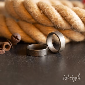 May include: Two silver-toned metal rings on a dark gray surface. The rings are simple and have a brushed finish. The rings are in focus, while the background is blurred.