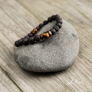 Puede incluir: Un brazalete de cuentas de lava negra y ojo de tigre sobre una piedra gris. El brazalete tiene cuentas de madera intercaladas con las cuentas de lava y ojo de tigre.