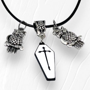 May include: A black cord necklace featuring three silver-toned charms. Two owl charms flank a central charm with a white coffin pendant with a black cross design. The charms are detailed with intricate patterns.